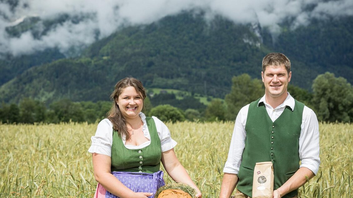 Ein Paar in traditioneller Tracht steht vor einer malerischen Berglandschaft. Sie halten ein Holzfass und eine Tüte Mehl in der Hand, umgeben von einem Feld. | © Netzwerk Kulinarik wildbild