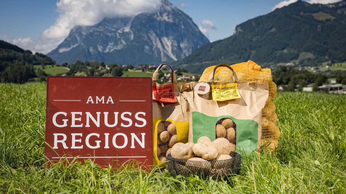 Ein Zeichen der AMA Genuss Region steht vor einer malerischen Berglandschaft. Im Vordergrund sind Taschen mit Kartoffeln und anderen landwirtschaftlichen Produkten zu sehen. | © Netzwerk Kulinarik Wildbild