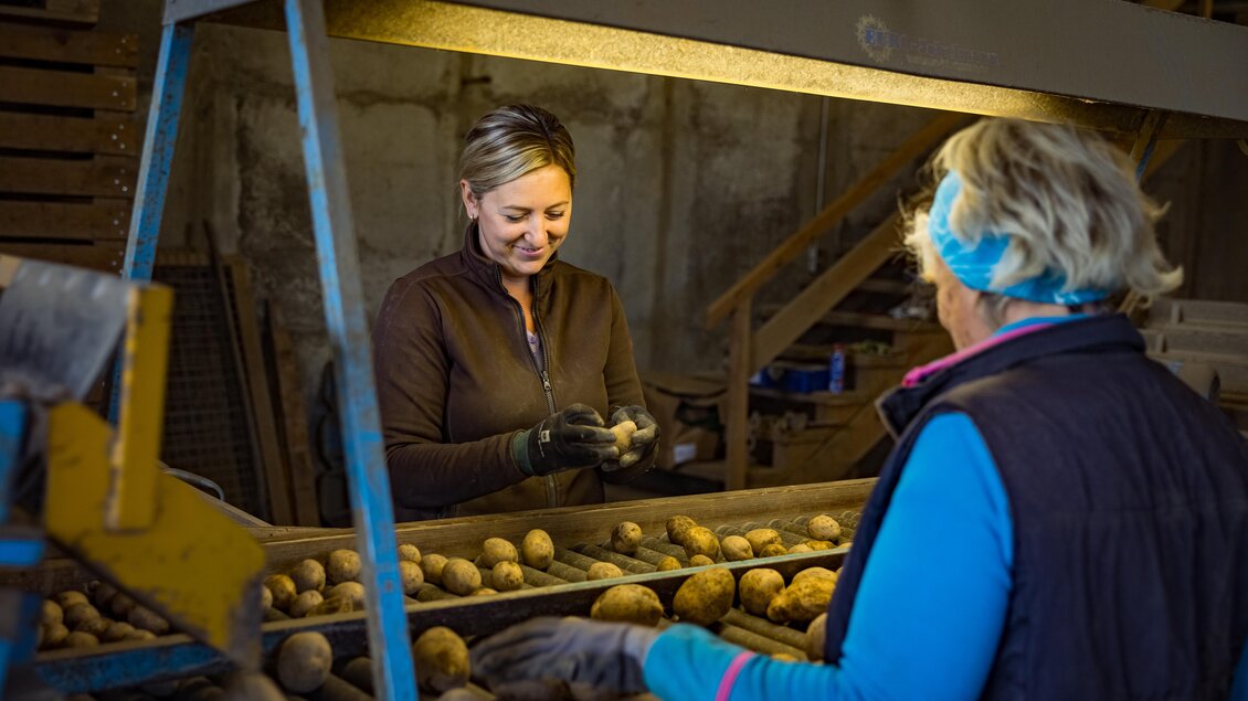 Zwei Frauen arbeiten an einer Kartoffelverarbeitungsmaschine. Eine Frau sortiert die Kartoffeln, während die andere lächelt und hilft. | © Netzwerk Kulinarik Wildbild