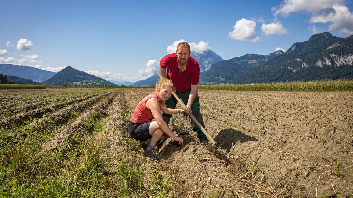 Zwei Personen arbeiten auf einem Feld und bereiten den Boden vor. Im Hintergrund sind Berge und ein blauer Himmel zu sehen. | © Netzwerk Kulinarik Wildbild