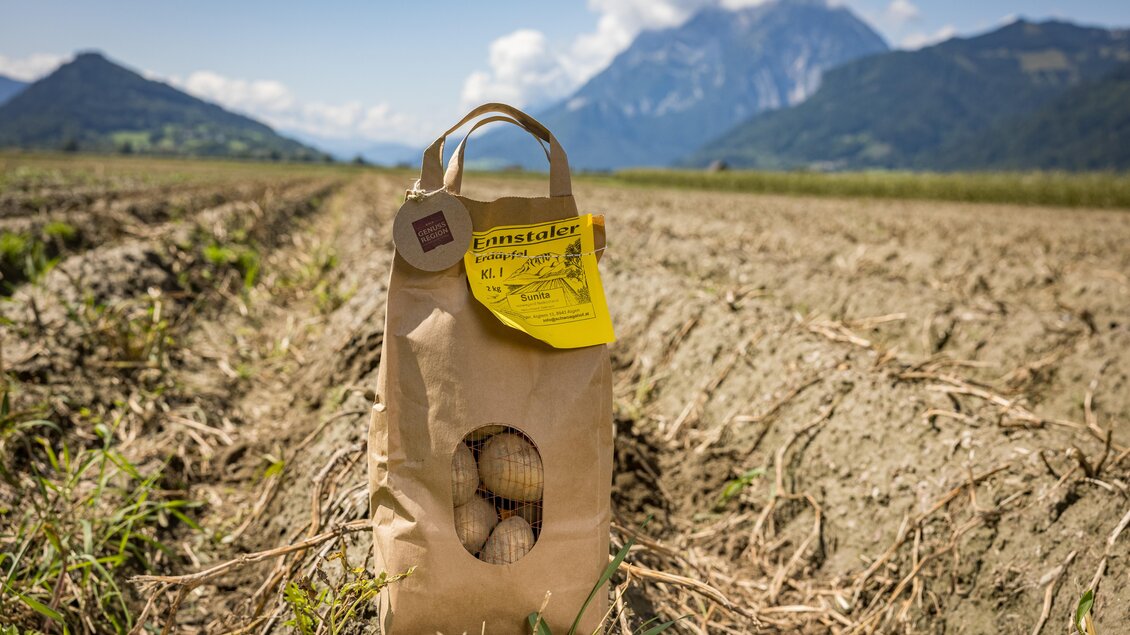 Ein Papiertüte mit Eiern steht auf einem unbewirtschafteten Feld. Im Hintergrund sind Berge und ein blauer Himmel zu sehen. | © Netzwerk Kulinarik Wildbild