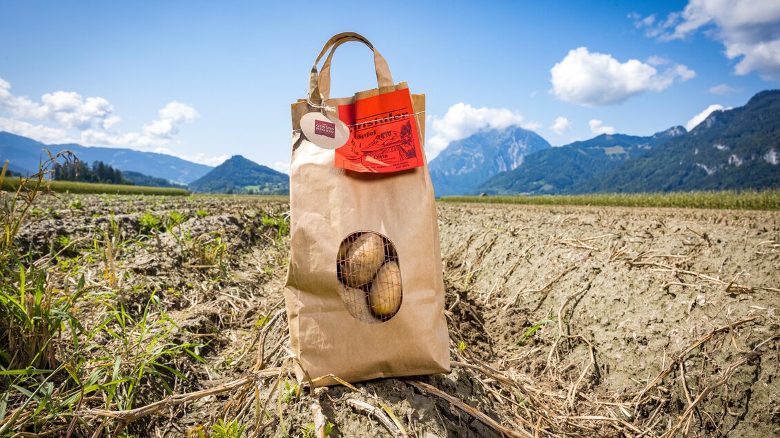 Eine braune Papiertüte steht auf einem Feld, die mit drei fertigen Kartoffeln gefüllt ist. Im Hintergrund sind Berge und ein blauer Himmel mit wenigen Wolken zu sehen. | © Netzwerk Kulinarik Wildbild