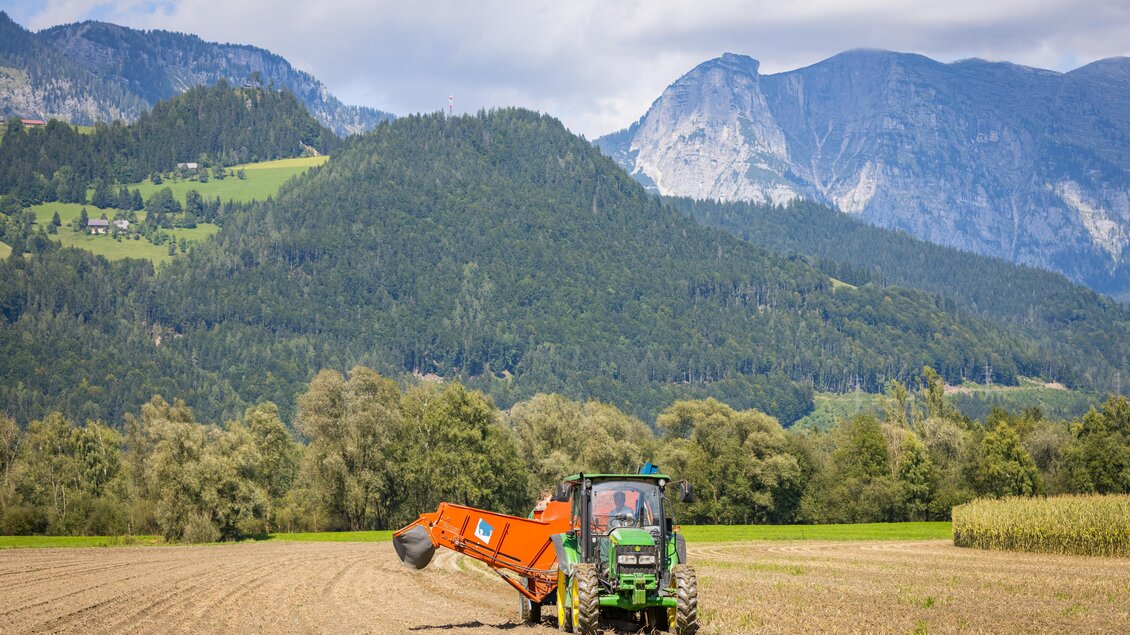 Ein Traktor arbeitet auf einem Feld mit den Bergen im Hintergrund. Der Himmel ist teilweise bewölkt und die umliegende Landschaft ist grün und hügelig. | © Netzwerk Kulinarik wildbild