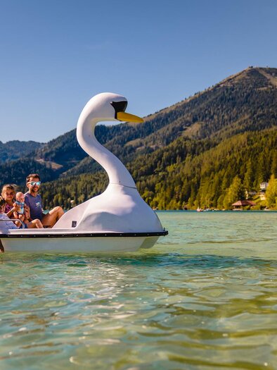 Eine Familie fährt mit einem Schwansanboot auf einem klaren See. Im Hintergrund sind grüne Hügel und Bäume zu sehen. | © TV Hochsteiermark | Rudy Dellinger