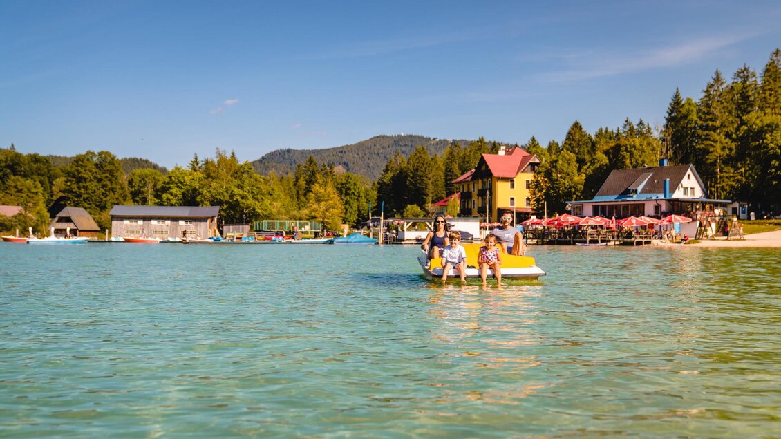 Eine Gruppe von Menschen paddelt auf einem Wassergefährt in klarem, blauem Wasser. Im Hintergrund sind schöne Häuser und Bäume zu sehen. | © TV Hochsteiermark | Rudy Dellinger
