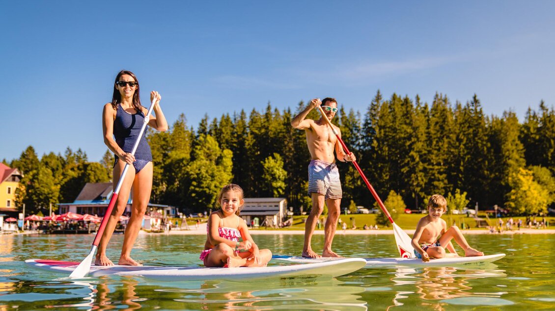 Eine Familie steht auf Stand-Up-Paddleboards im klaren Wasser. Im Hintergrund sind Bäume und Ferienhäuser sichtbar. | © TV Hochsteiermark | Rudy Dellinger
