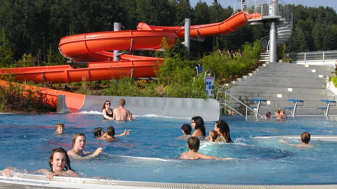 Ein Wasserpark mit orangefarbenen Rutschen und vielen schwimmenden Menschen im Pool. Die Umgebung ist grün und einladend. | © Erlebnisbad Eibiswald