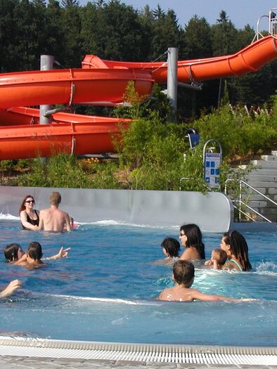 Ein Wasserpark mit orangefarbenen Rutschen und vielen schwimmenden Menschen im Pool. Die Umgebung ist grün und einladend. | © Erlebnisbad Eibiswald