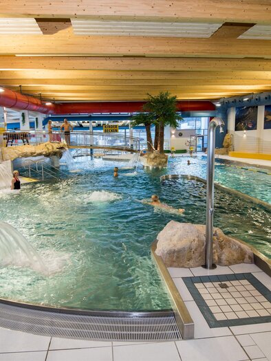 The photo shows a large, brightly lit indoor swimming pool with several pools and water attractions.<br/>In the foreground, a strong jet of water flows from a curved stainless steel pipe into the round pool. Several people are swimming in the water. In the center of the image, there is an artificial rock landscape with a small bridge and a palm tree.<br/>Behind it, there is a sign reading "Children's Pool." The pool is surrounded by light tiles with yellow accents and rock decorations. The ceiling is made of light wooden panels, and large red and blue pipes run along the ceiling. In the background, another area with loungers can be seen.<br/></p> | © Shooting Star