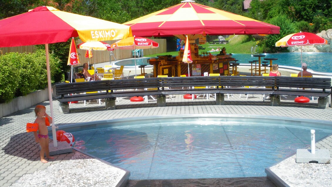 Ein Schwimmbad mit Sonnenschirmen und Liegen. Im Hintergrund ist ein gemütliches Café zu sehen. | © Marktgemeinde Stainach-Pürgg