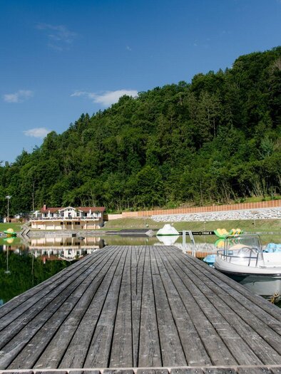 A beautiful lake with a wooden pier and boats. In the background, a wooded shoreline landscape can be seen. | © TV Region Graz - Mias Photoart