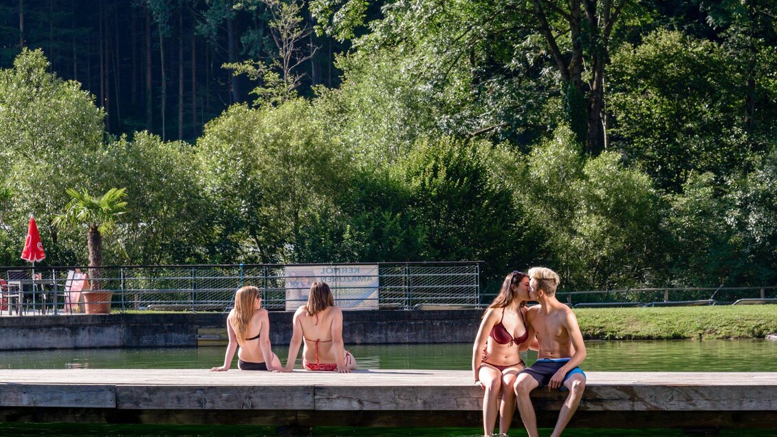 Zwei Paare sitzen am Ufer eines Sees und genießen die Sonne. Im Hintergrund sind grüne Bäume und eine ruhige Atmosphäre zu sehen. | © TV Region Graz - Mias Photoart
