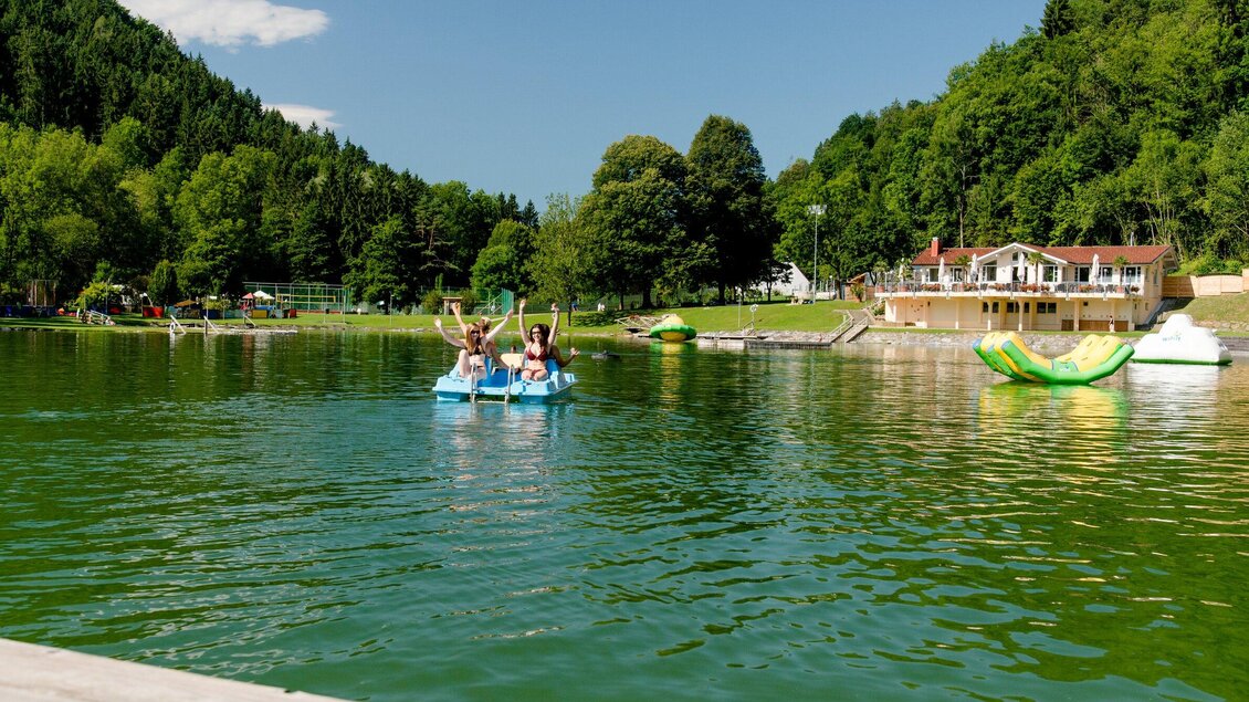 Ein ruhiger See umgeben von Bäumen und einer schönen Landschaft. Zwei Personen paddeln fröhlich in einem Tretboot. | © TV Region Graz - Mias Photoart