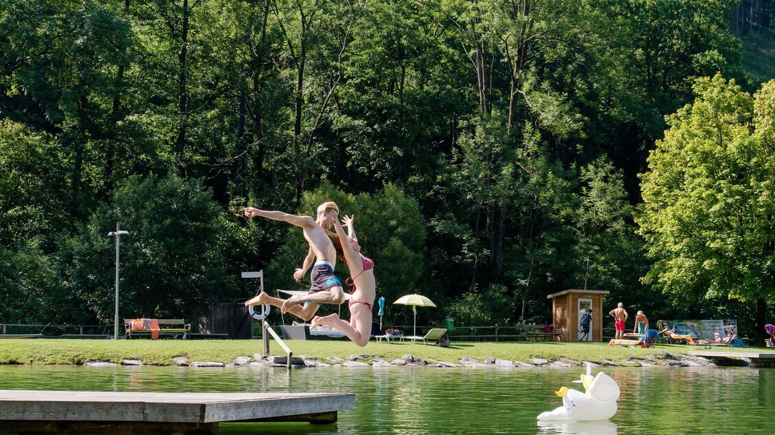 Zwei Kinder springen fröhlich von einem Steg ins Wasser. Im Hintergrund sind Bäume und weitere Menschen am See zu sehen. | © TV Region Graz - Mias Photoart