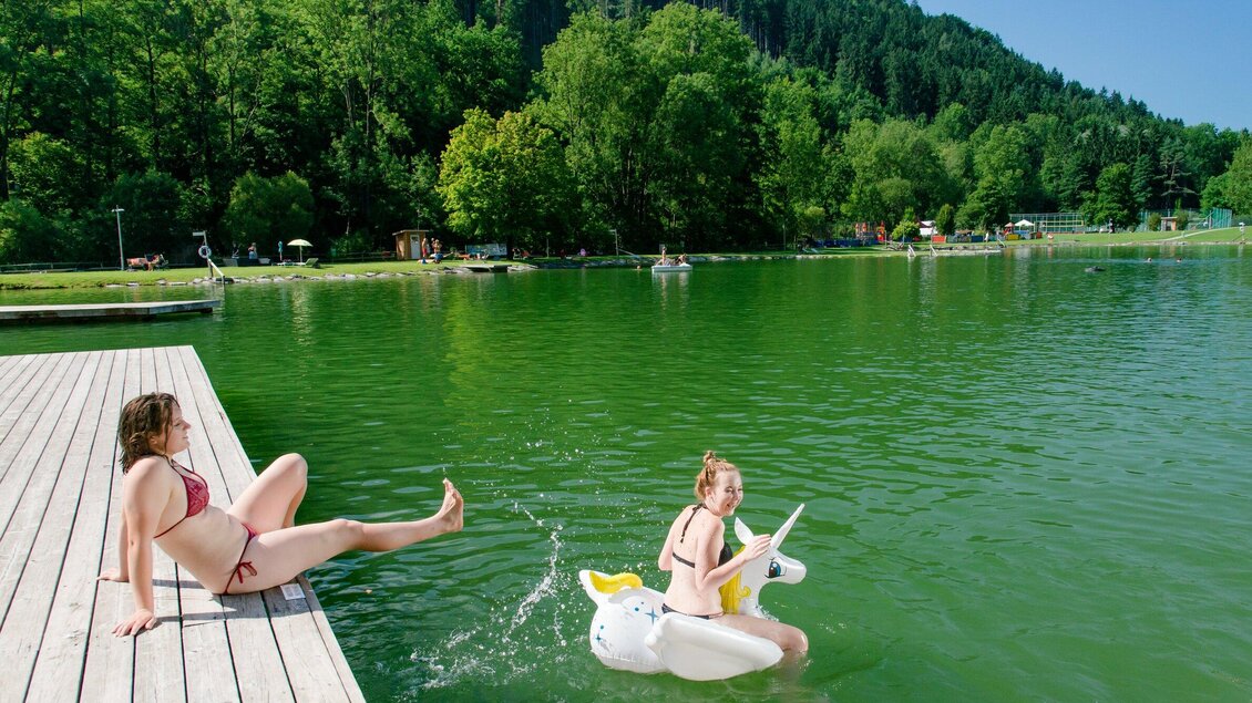 Zwei Frauen am Wasser. Eine entspannt auf einem Steg, während die andere auf einem aufblasbaren Einhorn schwimmt. | © TV Region Graz - Mias Photoart