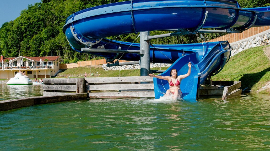 Eine Frau rutscht von einer blauen Wasserrutsche ins Wasser. Im Hintergrund sind grüne Bäume und einige Gebäude sichtbar. | © TV Region Graz - Mias Photoart