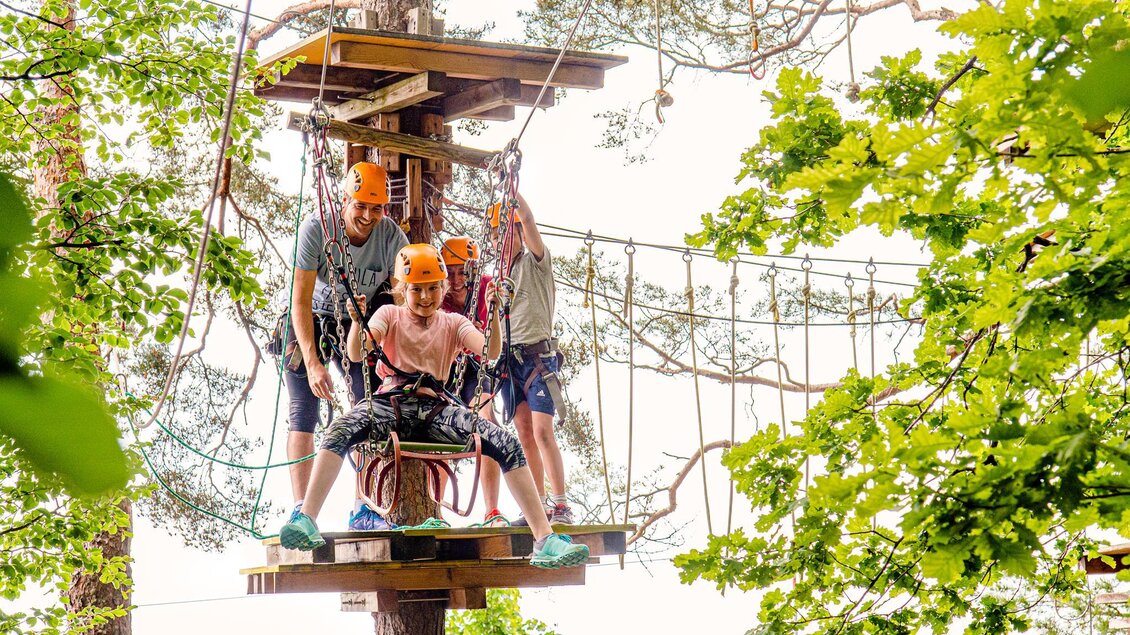 Drei Personen auf einer Plattform im Hochseilgarten, bereit für eine Herausforderung. Bäume und grüne Blätter umgeben die Szene. | © Miasphotoart
