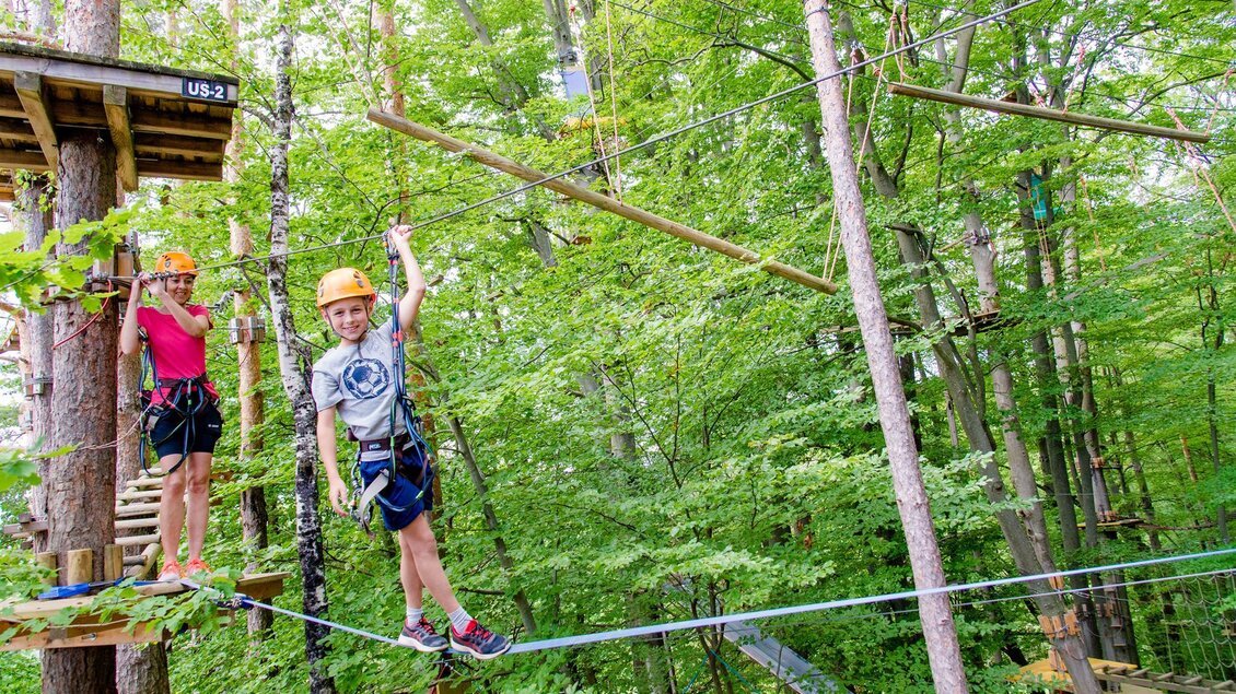 Zwei Kinder in einem Hochseilgarten zwischen Bäumen. Sie tragen Helme und klettern über Seile in der grünen Umgebung. | © Mias Photoart