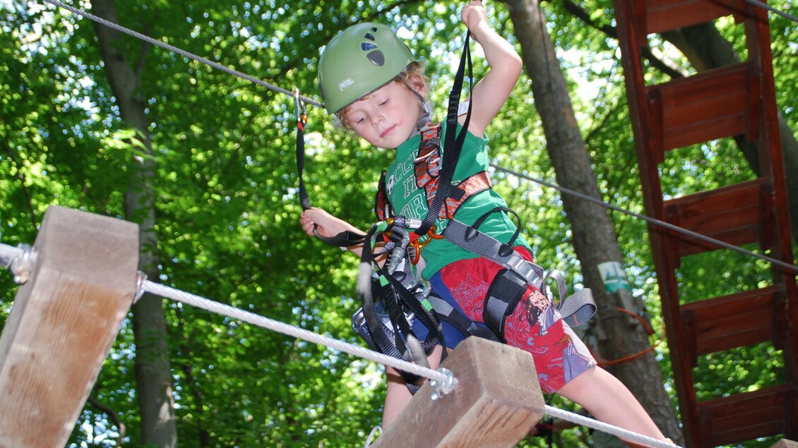Ein Junge klettert an einem Hochseilgarten. Er trägt einen Helm und Sicherheitsgeschirr zwischen Bäumen. | © Schnurr