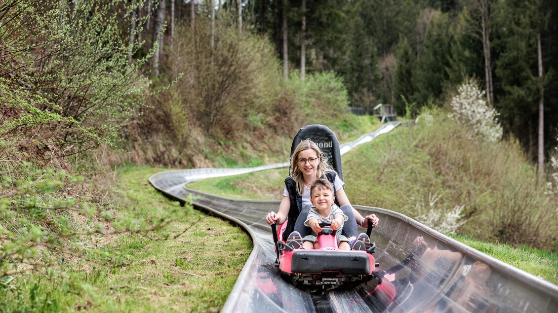 Eine Frau und ein Kind fahren gemeinsam eine Sommerrodelbahn. Umgeben von Bäumen und grünem Gras erleben sie Spaß und Abenteuer. | © Tourismusverband Oststeiermark