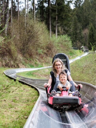 A woman and a child are riding a summer toboggan run together. Surrounded by trees and green grass, they are experiencing fun and adventure. | © Tourismusverband Oststeiermark