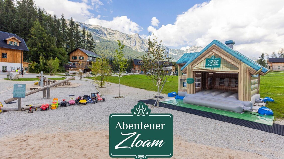 Ein schöner Spielplatz mit einem großen Hüpfhaus und verschiedenen Spielzeugen. Im Hintergrund sind Holzhütten und Berge zu sehen. | © www.zloam.at