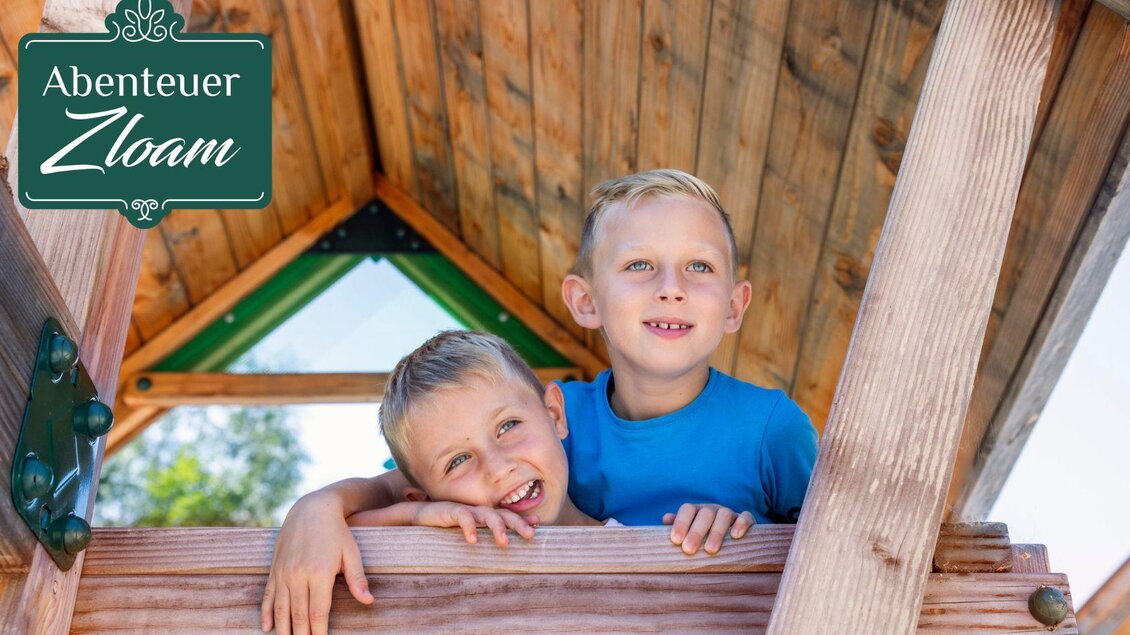 Zwei fröhliche Jungen stehen in einer Holzhütte und lächeln in die Kamera. Der Hintergrund zeigt einen klaren Himmel und grüne Bäume. | © www.zloam.at