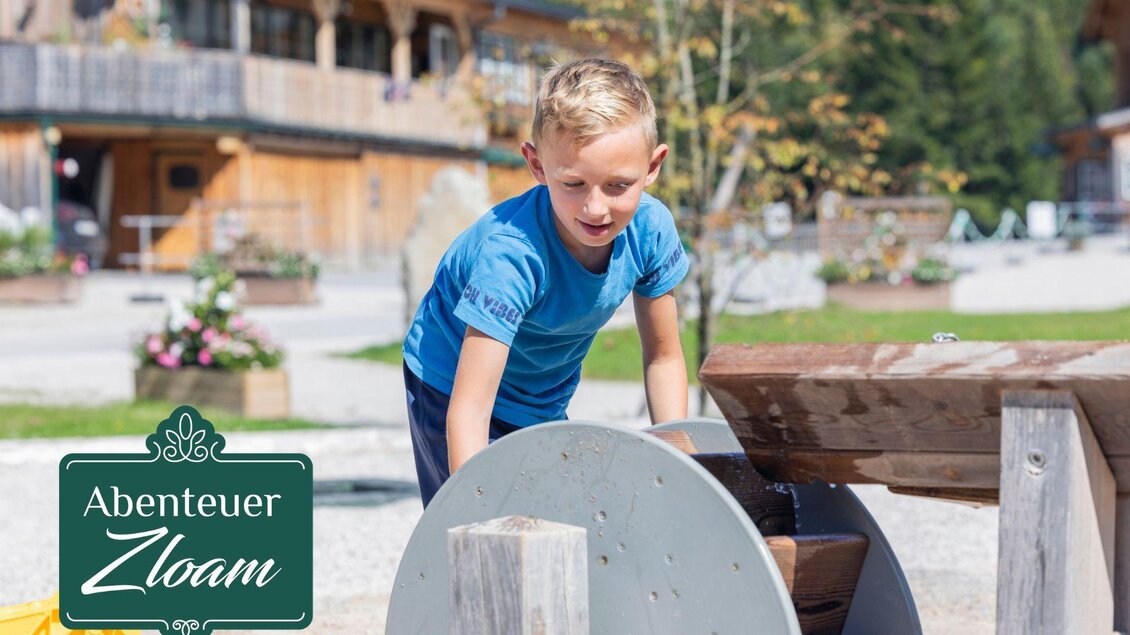 Ein Junge spielt mit einem Wasserlauf an einem Spielplatz. Im Hintergrund sind Holzhäuser und Pflanzen zu sehen. | © www.zloam.at
