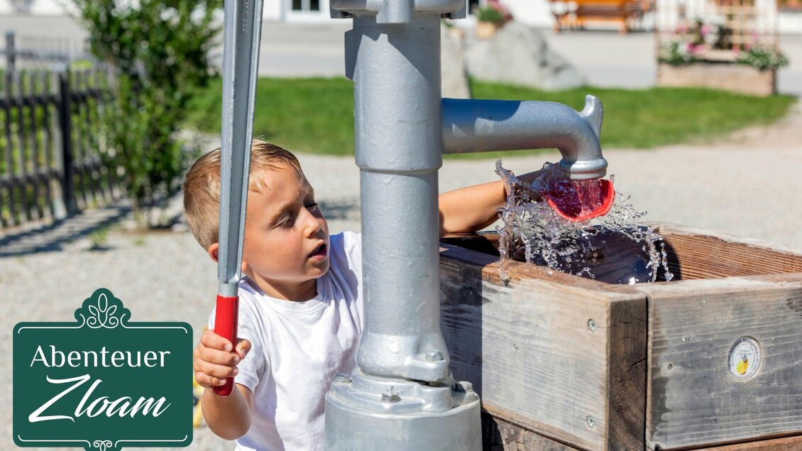 Ein Junge spielt an einer Wasserpumpe und spritzt Wasser. Im Hintergrund sind Grünflächen und Gebäude zu sehen. | © www.zloam.at