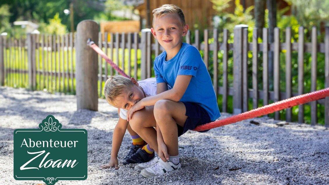 Zwei Kinder spielen auf einem Spielplatz. Eines sitzt auf einer Schaukel und das andere gräbt im Sand. | © www.zloam.at