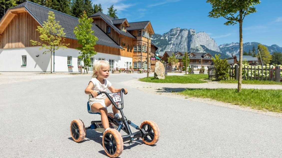 Ein kleines Kind fährt fröhlich auf einem Dreirad. Im Hintergrund sind hübsche Häuser und Berge zu sehen. | © www.zloam.at