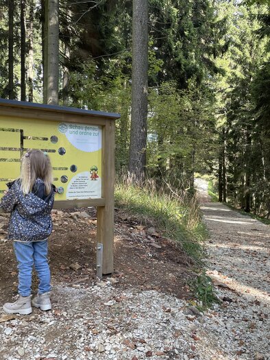 Ein Kind steht vor einem Informationsschild im Wald. Der Weg führt durch die Bäume und lädt zum Spazieren ein. | © Osteiermark Tourismus, Manuela Schnur