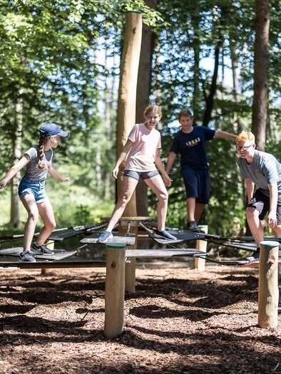 A group of children is playing on a playground in the forest. They are balancing on a wooden structure next to trees and enjoying the sunny day. | © Stadtmarketing Fürstenfeld