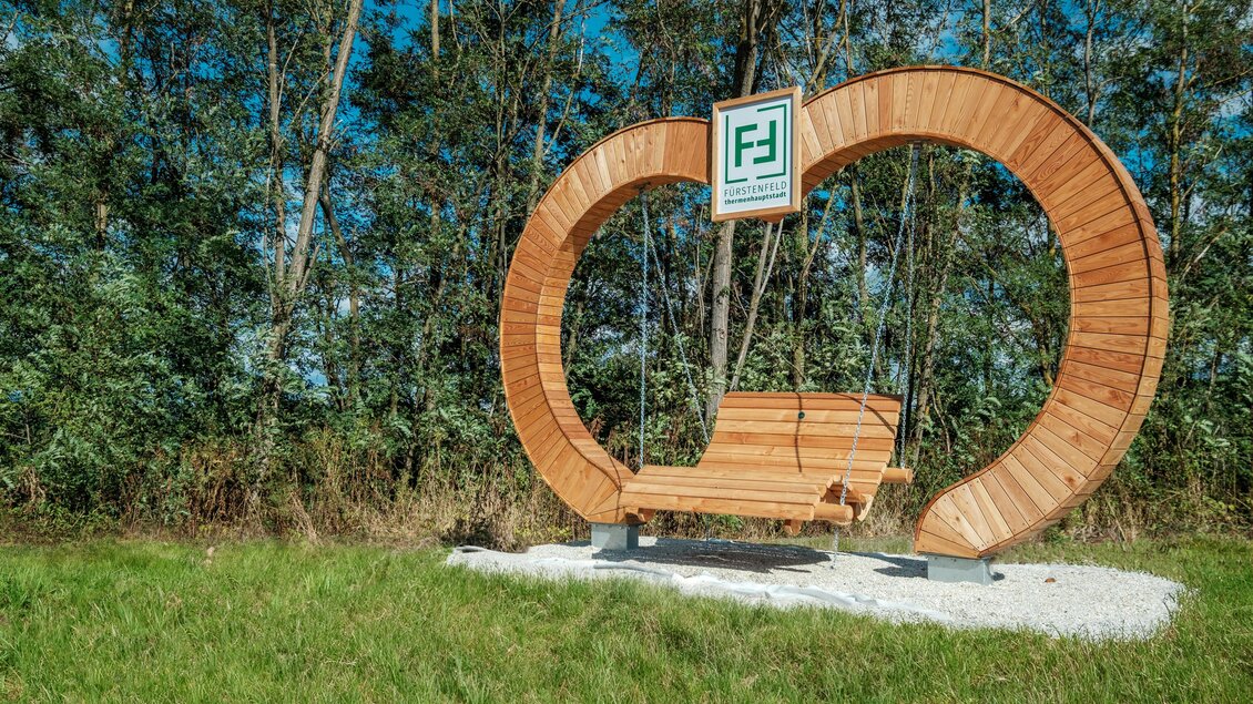 Eine hölzerne Schaukel in Herzform steht inmitten von Bäumen. Der Hintergrund zeigt grünes Gras und einen klaren Himmel. | © (c)Erlebniswelt-Fuerstenfeld_M-Effekt_Erlebniswelt