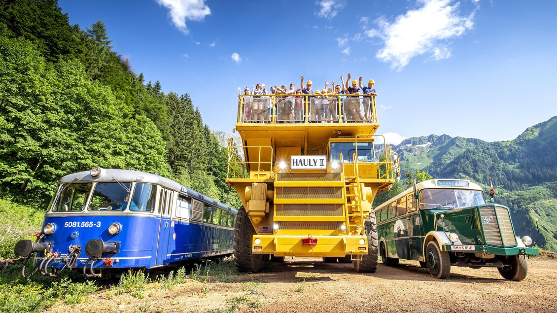 Ein blauer Zug und ein alter Bus stehen neben einem großen gelben Bagger. Im Hintergrund sind grüne Berge und ein blauer Himmel zu sehen. | © August Zoebl
