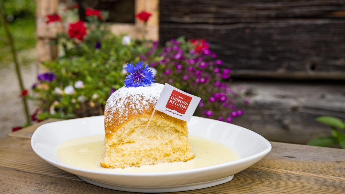 Ein Stück Kuchen mit Puderzucker und einer kleinen Fahne, serviert auf einer weißen Schale in einer cremigen Sauce. Im Hintergrund sind bunte Blumen zu sehen. | © Netzwerk Kulinarik wildbild