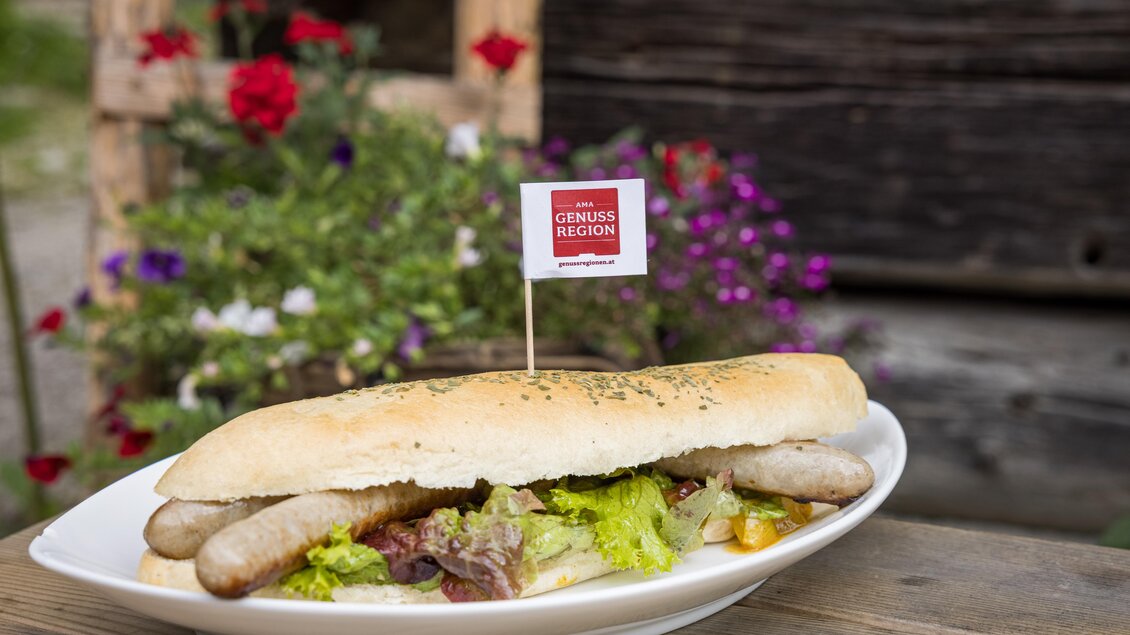 Ein Wurstbrötchen mit frischem Gemüse auf einem Teller. Im Hintergrund sind bunte Blumen zu sehen. | © Netzwerk Kulinarik wildbild