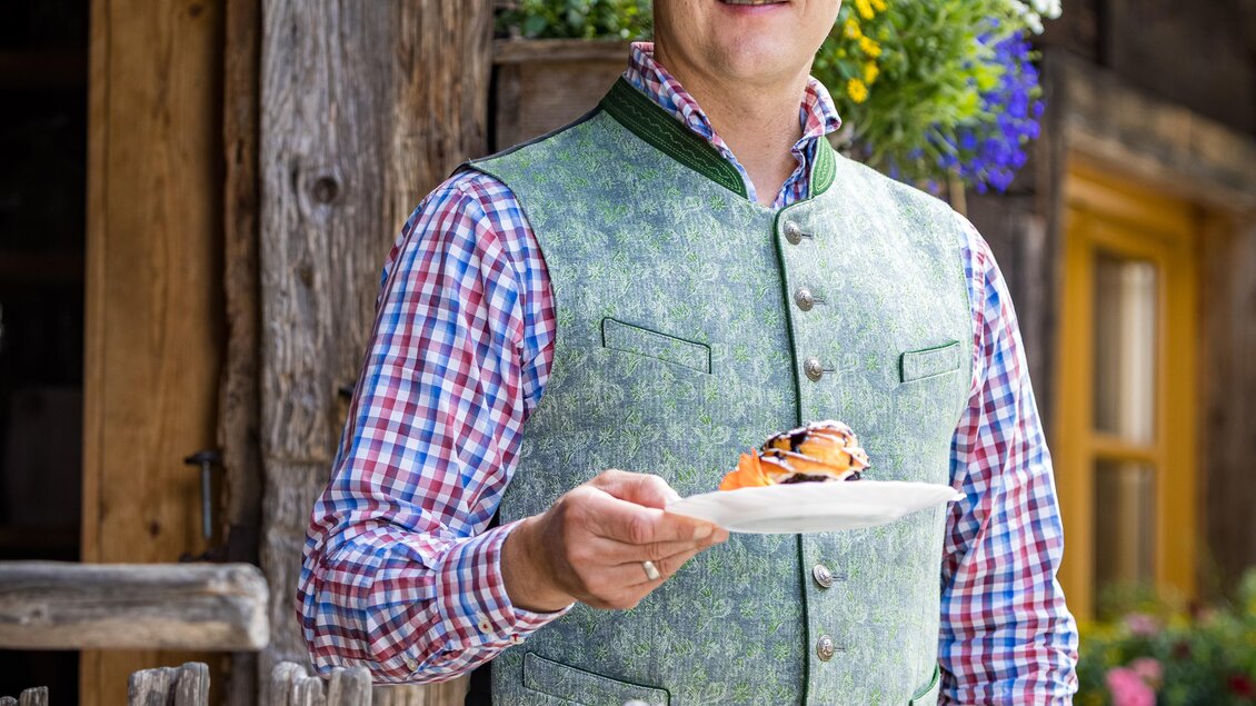 Ein Mann steht vor einem Holzhaus und hält einen Teller mit Essen in der Hand. Im Hintergrund sind Blumen und ein Holzzaun zu sehen. | © Netzwerk Kulinarik wildbild
