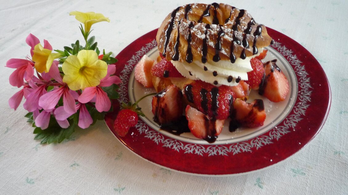 Ein köstliches Dessert mit frischen Erdbeeren, Sahne und einem Waffelstück, dekoriert mit Schokoladensauce. Neben dem Teller liegen bunte Blumen. | © Eschachalm