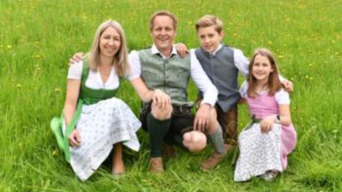 Eine Familie in traditioneller bayerischer Tracht sitzt auf einer Wiese. Im Hintergrund sind grüne Wiesen und Blumen zu sehen. | © Privat