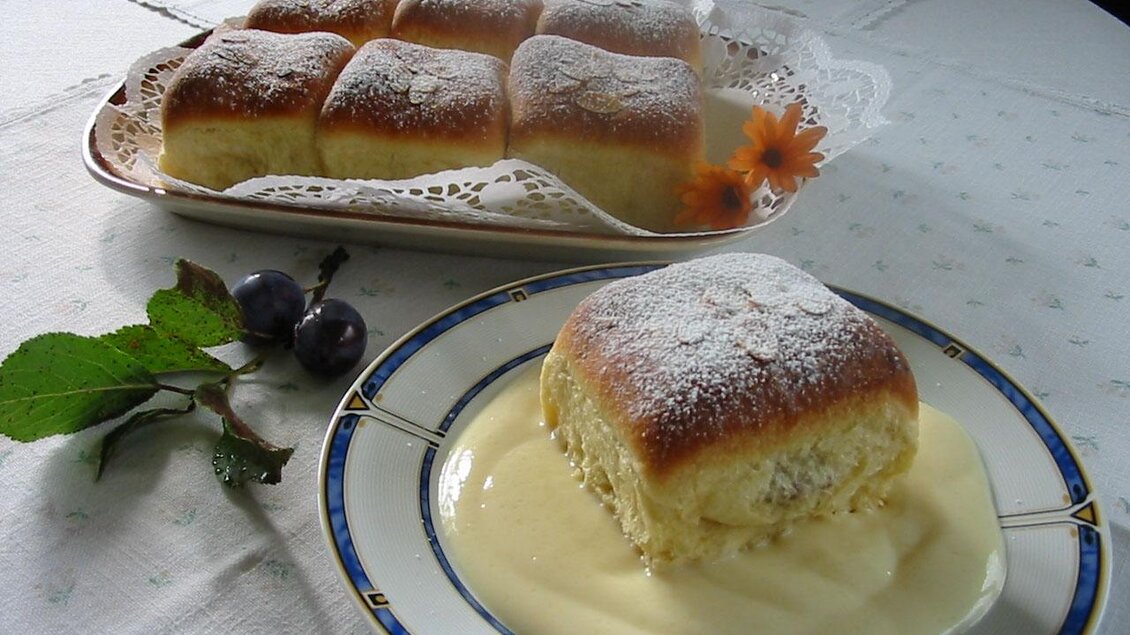 Ein Teller mit einem Stück süßem Gebäck, das mit Puderzucker bestäubt ist, liegt neben einer Schüssel mit cremiger Sauce. Im Hintergrund sind weitere Gebäckstücke auf einer Platten mit dekorativer Serviette. | © Eschachalm