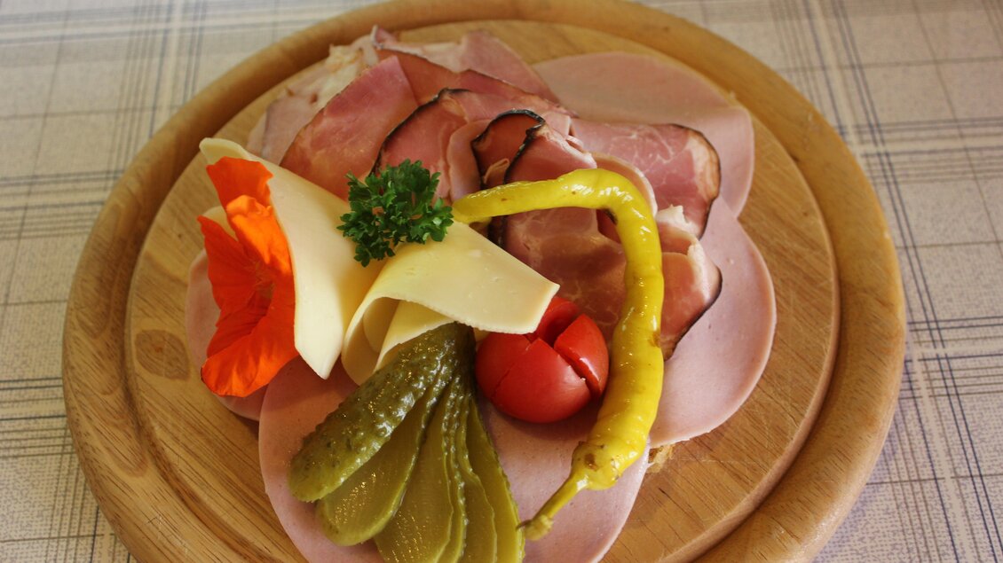 Eine appetitliche Wurstplatte mit verschiedenen Sorten Schinken, Käse und Essiggurken. Bunte Beilagen wie Tomate und eine essbare Blume runden das Bild ab. | © Eschachalm