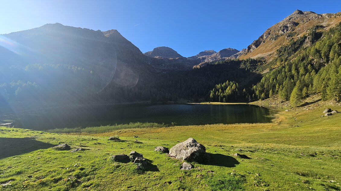 Eine malerische alpine Landschaft mit einem ruhigen See und grünen Wiesen. Im Hintergrund sind hohe Berge und blauer Himmel zu sehen. | © Eschachalm