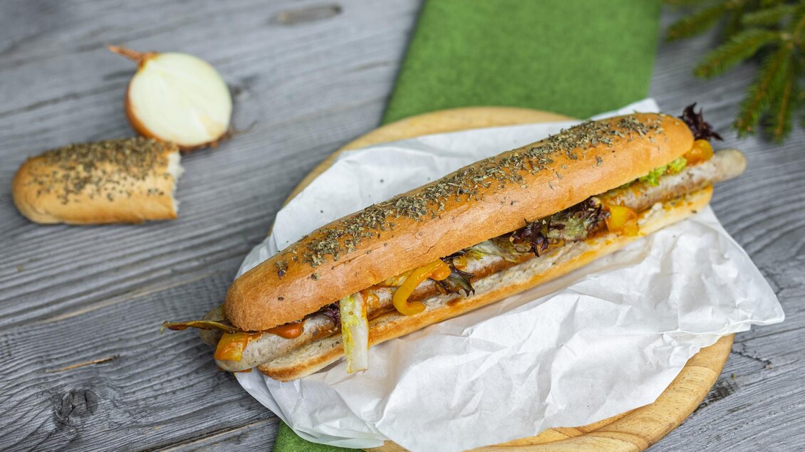 Ein belegtes Brötchen mit gegrilltem Fleisch, Zwiebeln und Käse auf einer Holzplatte. Im Hintergrund ist ein Stück Brot und eine grüne Unterlage zu sehen. | © Martin Huber