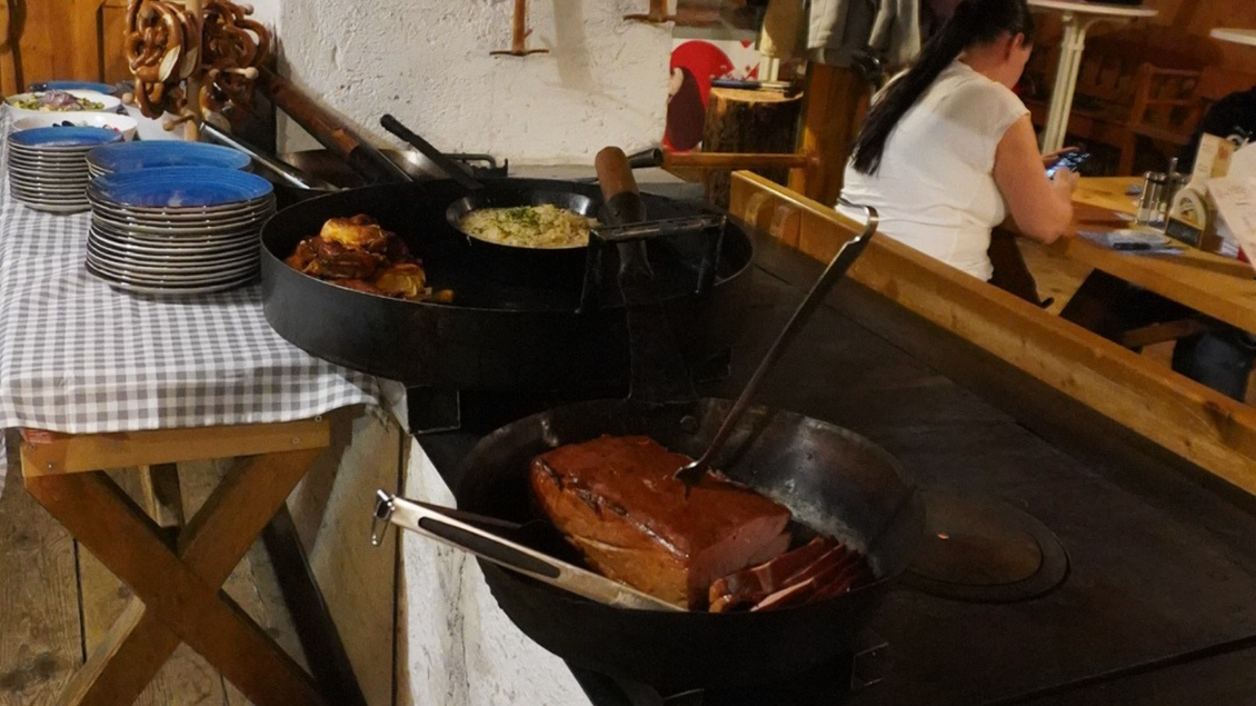 Ein rustikales Buffet mit verschiedenen warmen Gerichten in großen Pfannen. Im Hintergrund sieht man Gäste, die sich entspannen. | © Eselalm