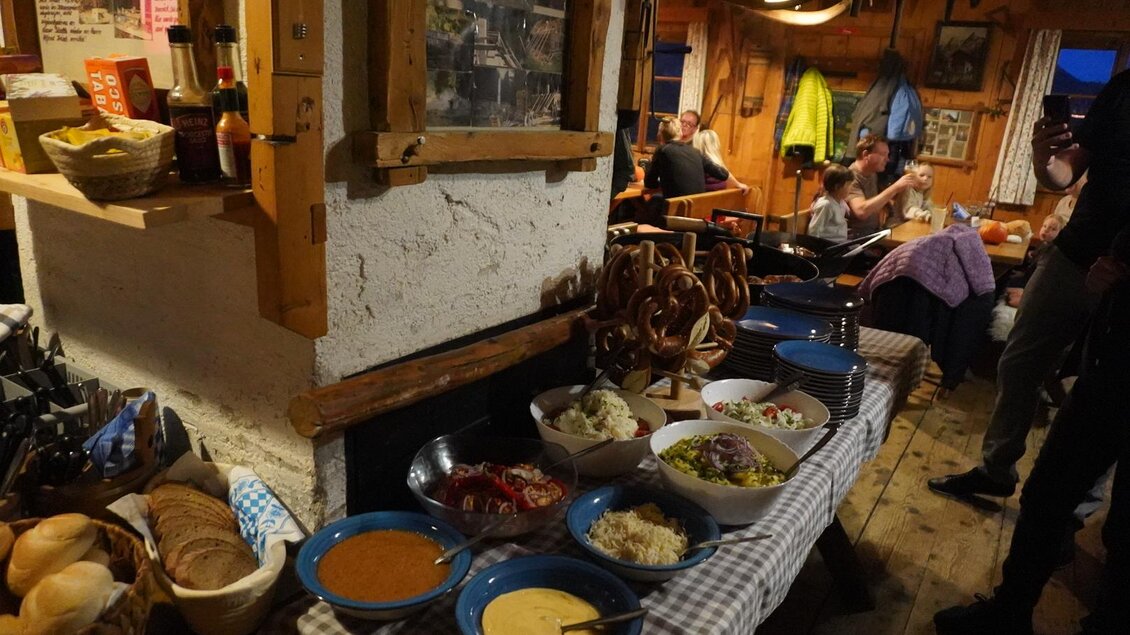 Ein rustikales Restaurant mit einem Tisch voller einladender Speisen, darunter Salate, Brot und Dips. Im Hintergrund sind Gäste zu sehen, die sich unterhalten. | © Eselalm