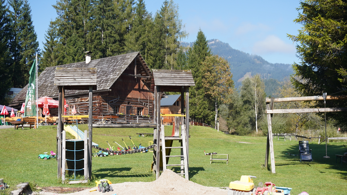Ein Spielplatz mit einer Rutsche und Schaukeln in einer grünen Wiese. Im Hintergrund steht ein Holzhaus umgeben von Bäumen und Bergen. | © Eselalm