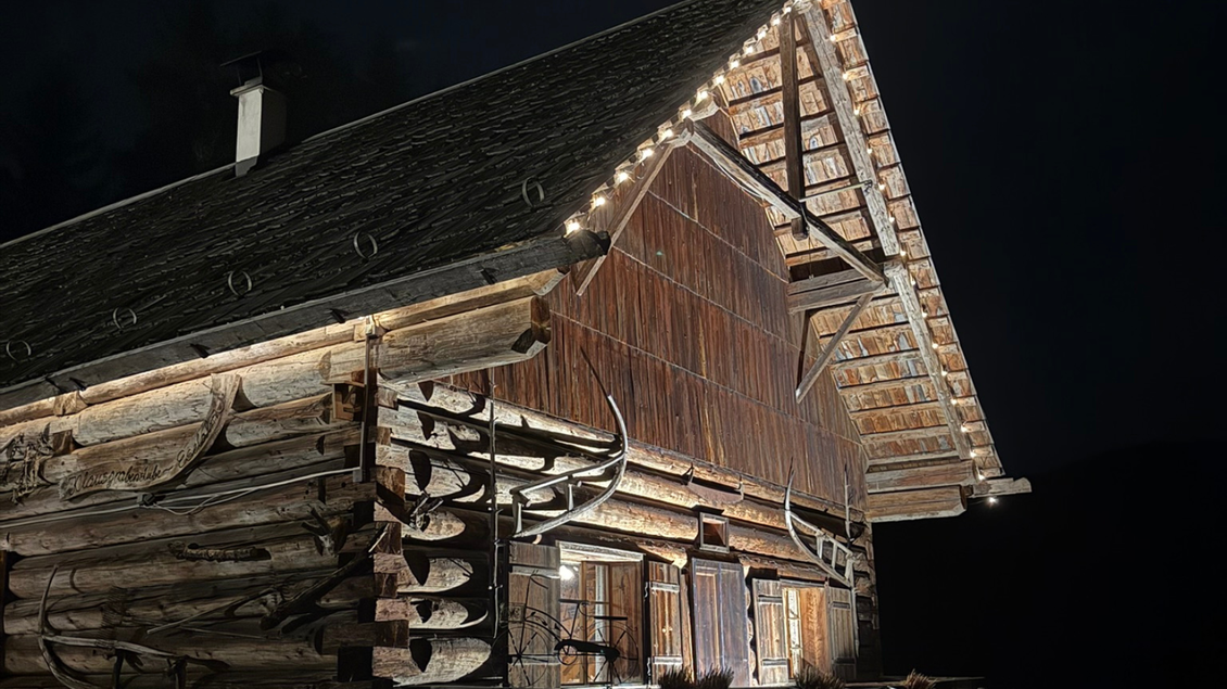 Ein rustikales Holzhaus, das in der Nacht beleuchtet ist. Die warmen Lichter heben die Struktur und Details des Gebäudes hervor. | © Eselalm