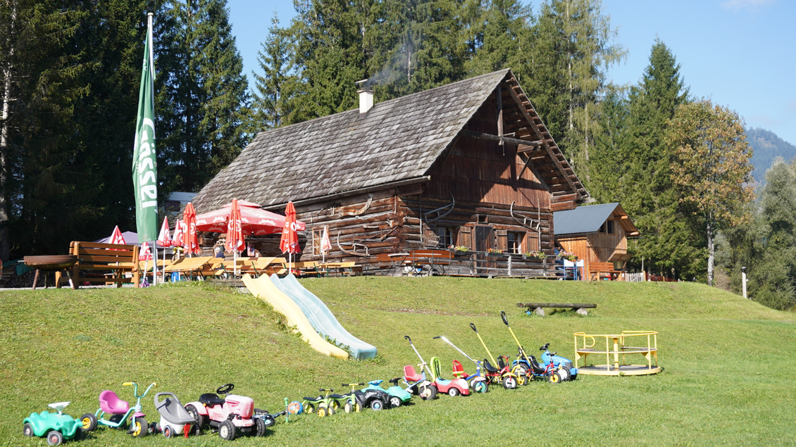 Ein Holzhaus in der Natur mit einem grünen Rasen. Vor dem Haus stehen viele Spielzeuge für Kinder, darunter Rutschbahnen und kleine Fahrzeuge. | © Eselalm