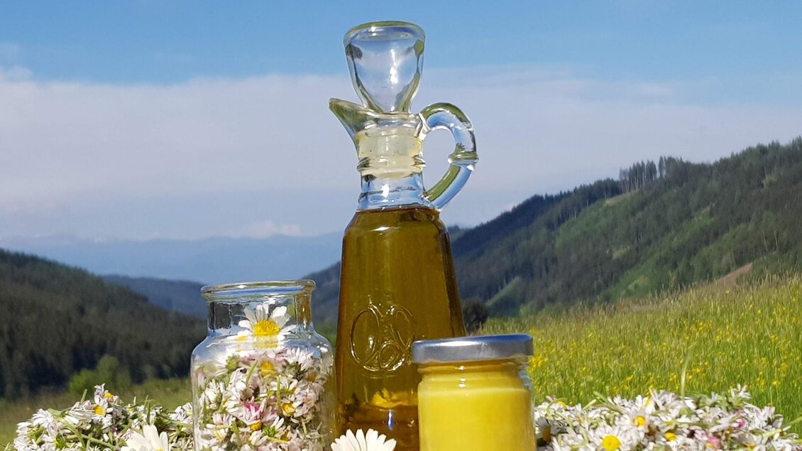 Ein Glas mit Olivenöl steht zusammen mit einem Honigglas und einer kleinen Schale, umgeben von Wiesenblumen. Im Hintergrund sind grüne Hügel und ein blauer Himmel zu sehen. | © Erlebnisregion Murtal